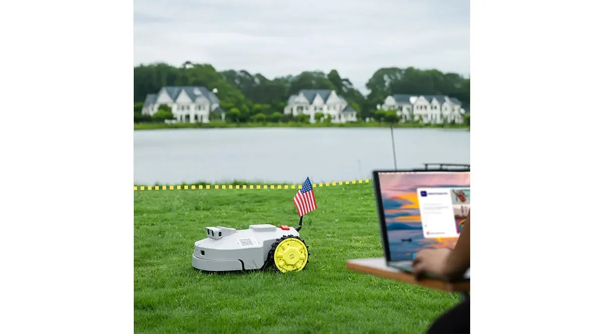 robotic lawn mower with American flag on green grass near lake, person working on laptop outdoors